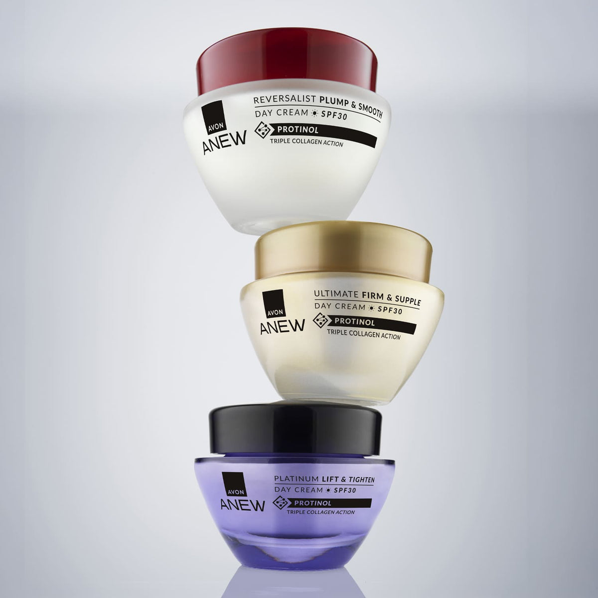 Three Anew skincare day cream jars stacked vertically on a light gray gradient background. The top jar has a red lid and white body, the middle has a gold lid and beige body, and the bottom has a black lid and purple body.