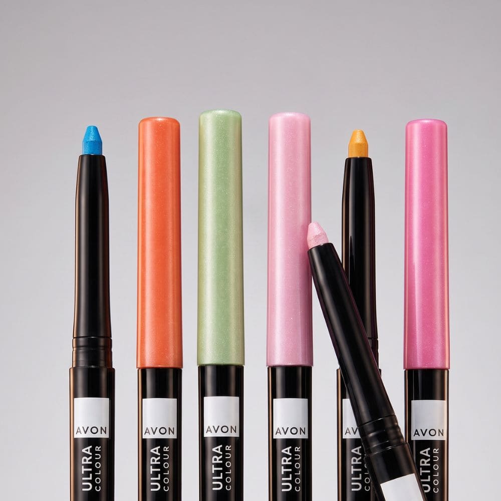 Six Avon Ultra Colour lip crayons in assorted shades are lined up against a neutral gray background, with one crayon lying diagonally in front.