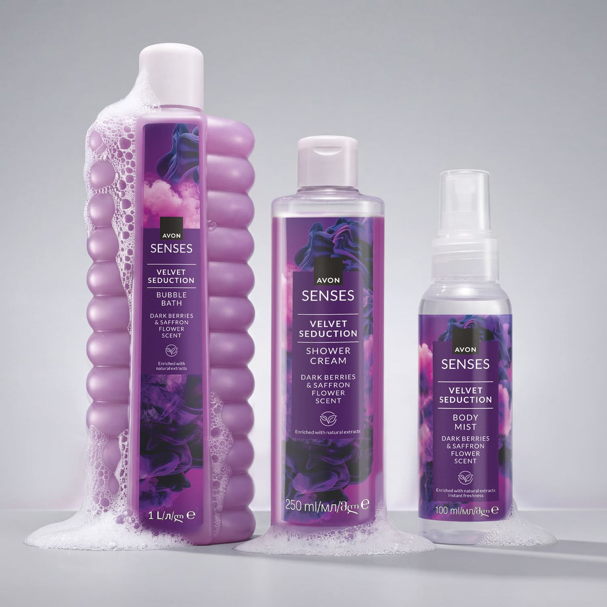 Three Avon Senses body care products in purple packaging arranged on a light gray background with white foam at the base: a large bubble bath bottle, a shower cream bottle, and a body mist spray bottle.