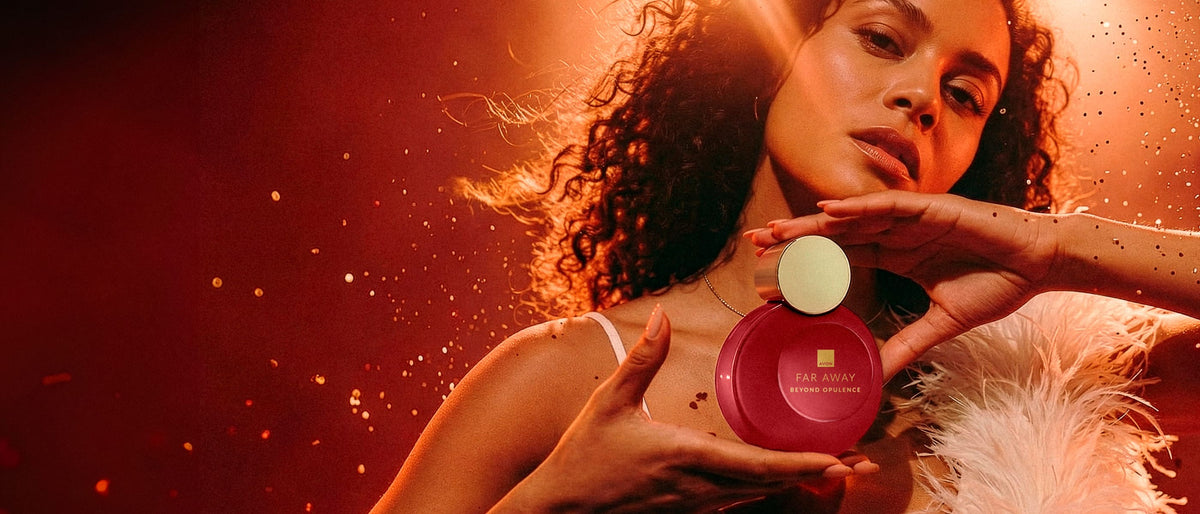 A woman with curly hair holds a round Far Away perfume bottle with a gold cap against a dramatic warm red-orange background with glowing particles.
