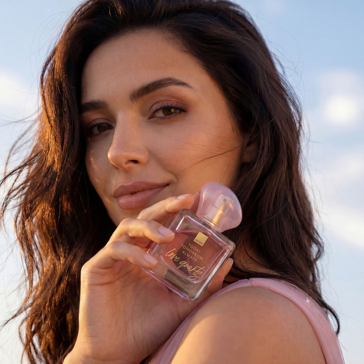 A woman outdoors holds a clear rectangular Avon perfume bottle with a translucent pink cap. The bottle has gold lettering that reads today, tomorrow, always.